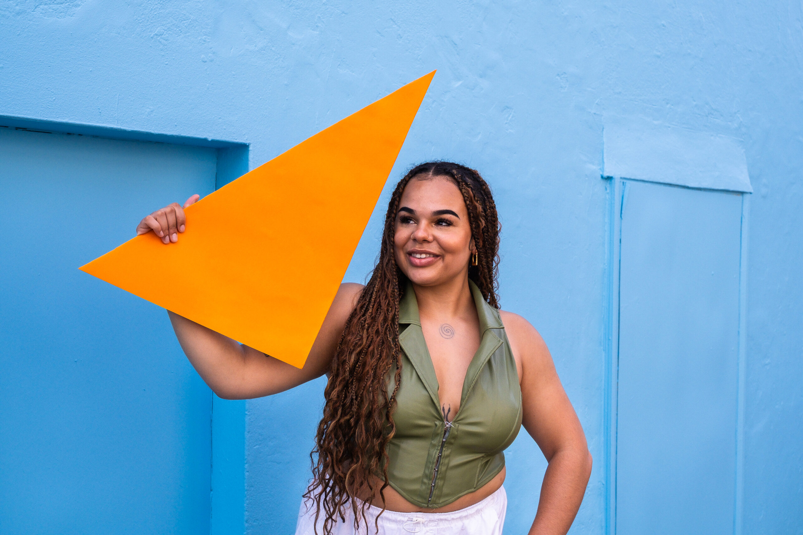 Woman with brown skin and dark long hair holding up an orange triangle against a blue wall