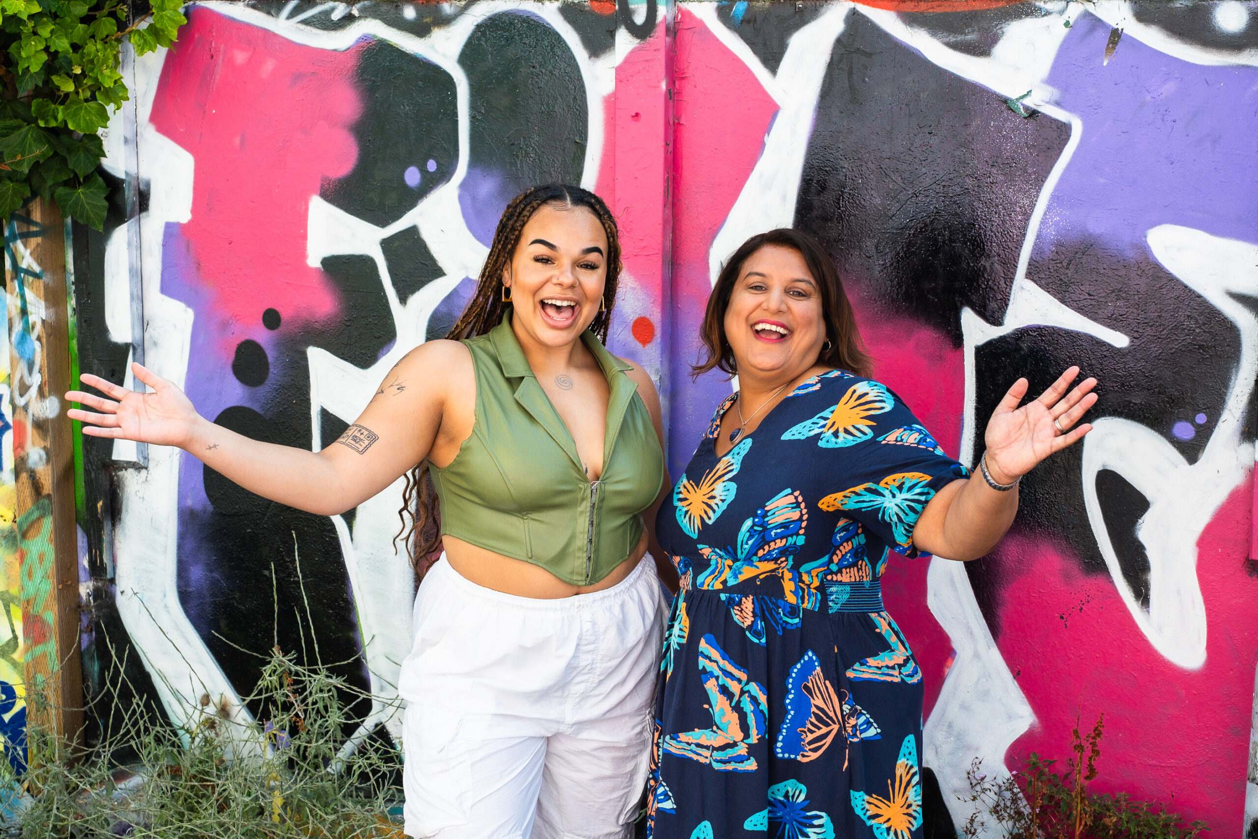 Mo and Kaia smiling with arms outstretched against a coloured wall