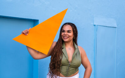 Woman with brown skin and dark long hair holding up an orange triangle against a blue wall