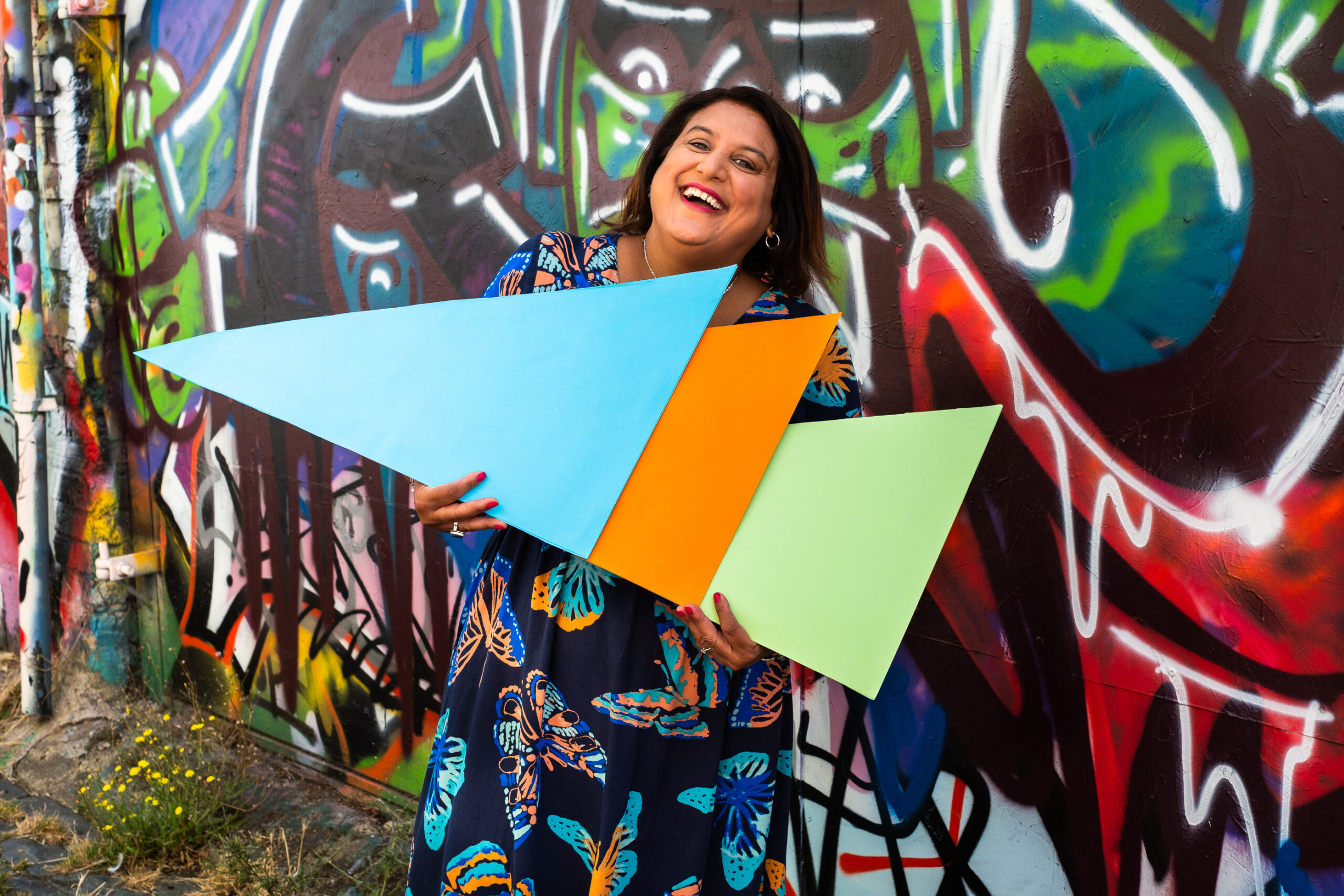 EOD-1019 Mo is holding three triangles, one blue, one orange and one light green, overlapping one another. She is smiling.