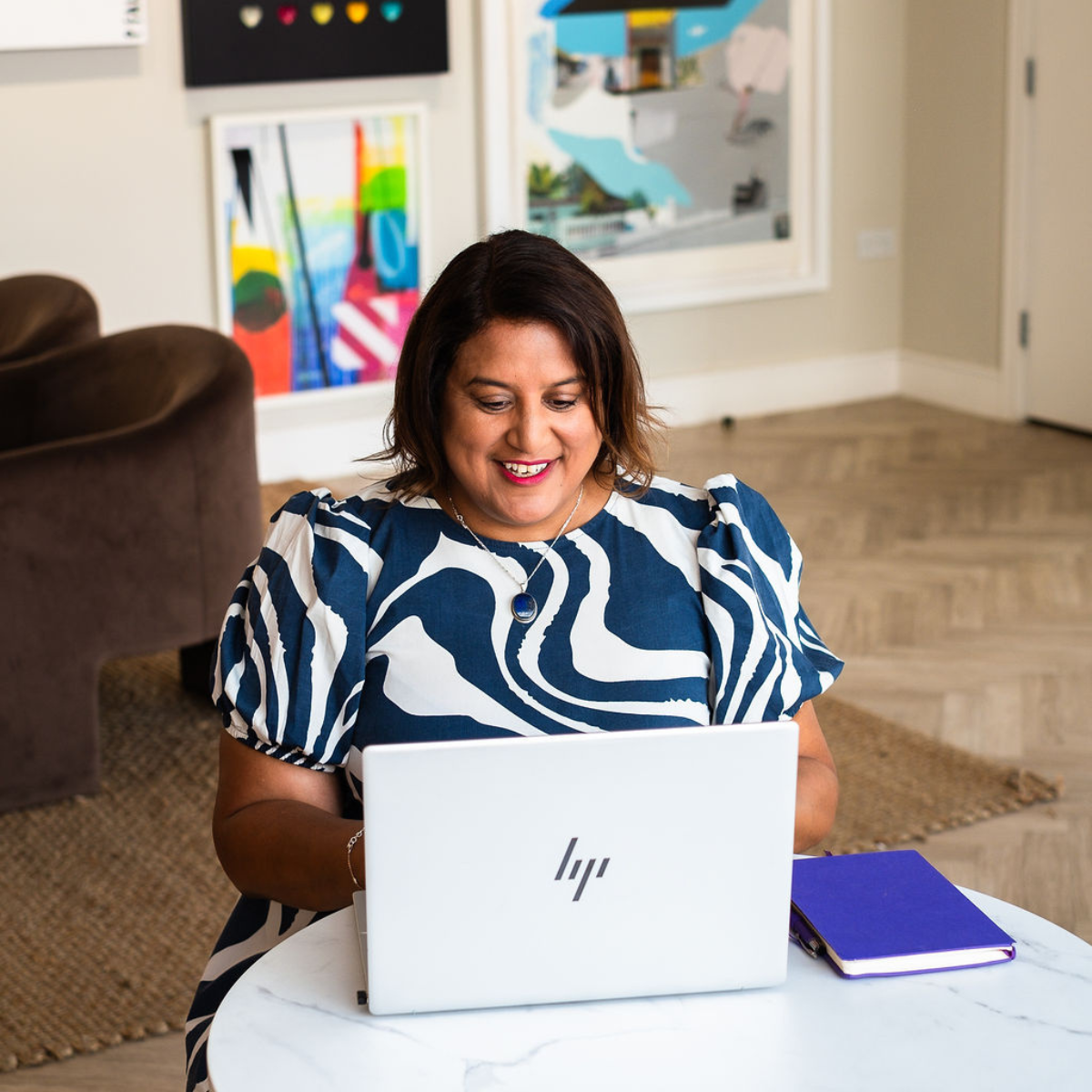 Mo is sitting at a circular desk, typing on a laptop with a purple notebook to her right. She is smiling, wearing a stripey patterned dress that is navy and white.