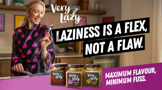 Advertisement for Very Lazy, a food brand. A smiling woman in a black kimono printed with pink garlic illustrations stands in a kitchen holding kitchen tongs. Three jars of Very Lazy chopped garlic, garlic and ginger, and chopped ginger are displayed in the foreground. Bold text reads: Laziness is a flex, not a flaw. Maximum flavour, minimum fuss.
